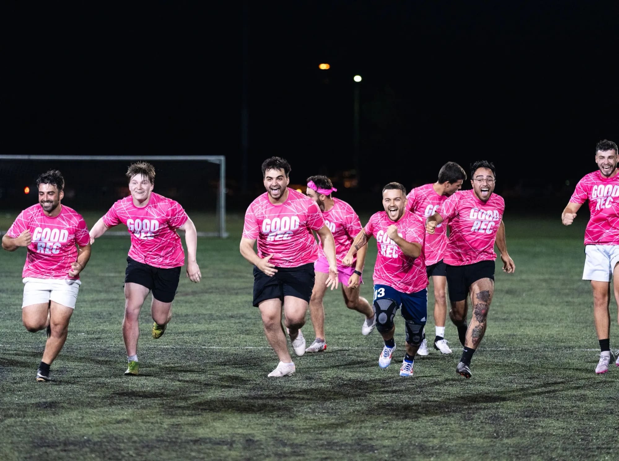 A team in GoodRec shirts running together in celebration across a floodlit field