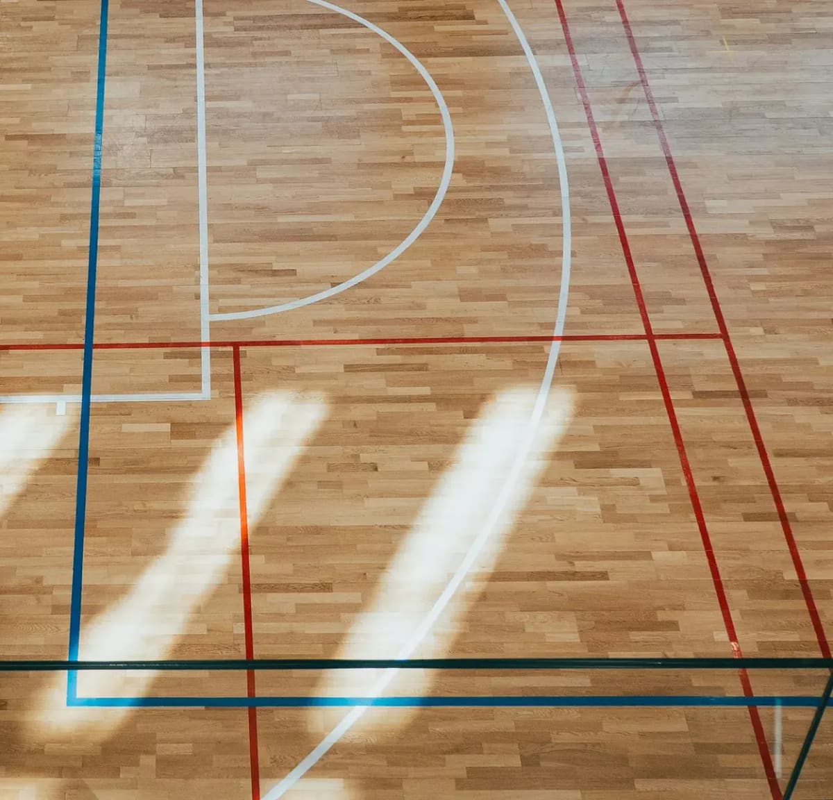 An empty indoor court seen from above, its lines crossing the wooden floor in the sun