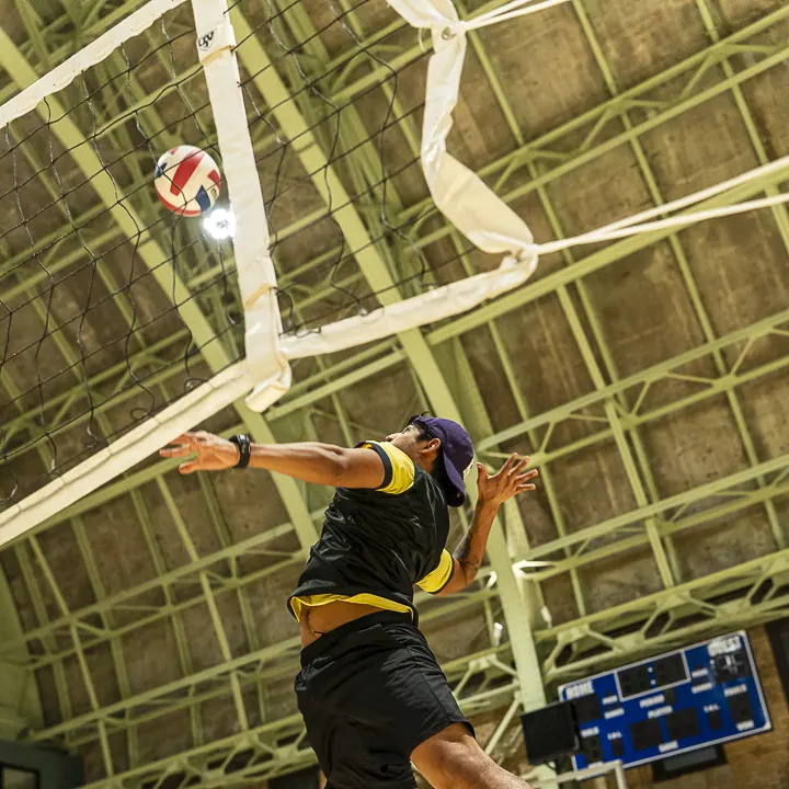 A player in a GoodRec bib leaping to spike indoors, the ball above the net
