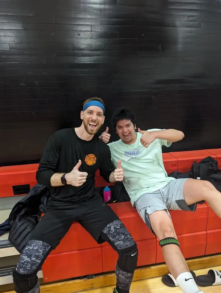 Two players grinning and giving a thumbs up on the bench of an indoor court