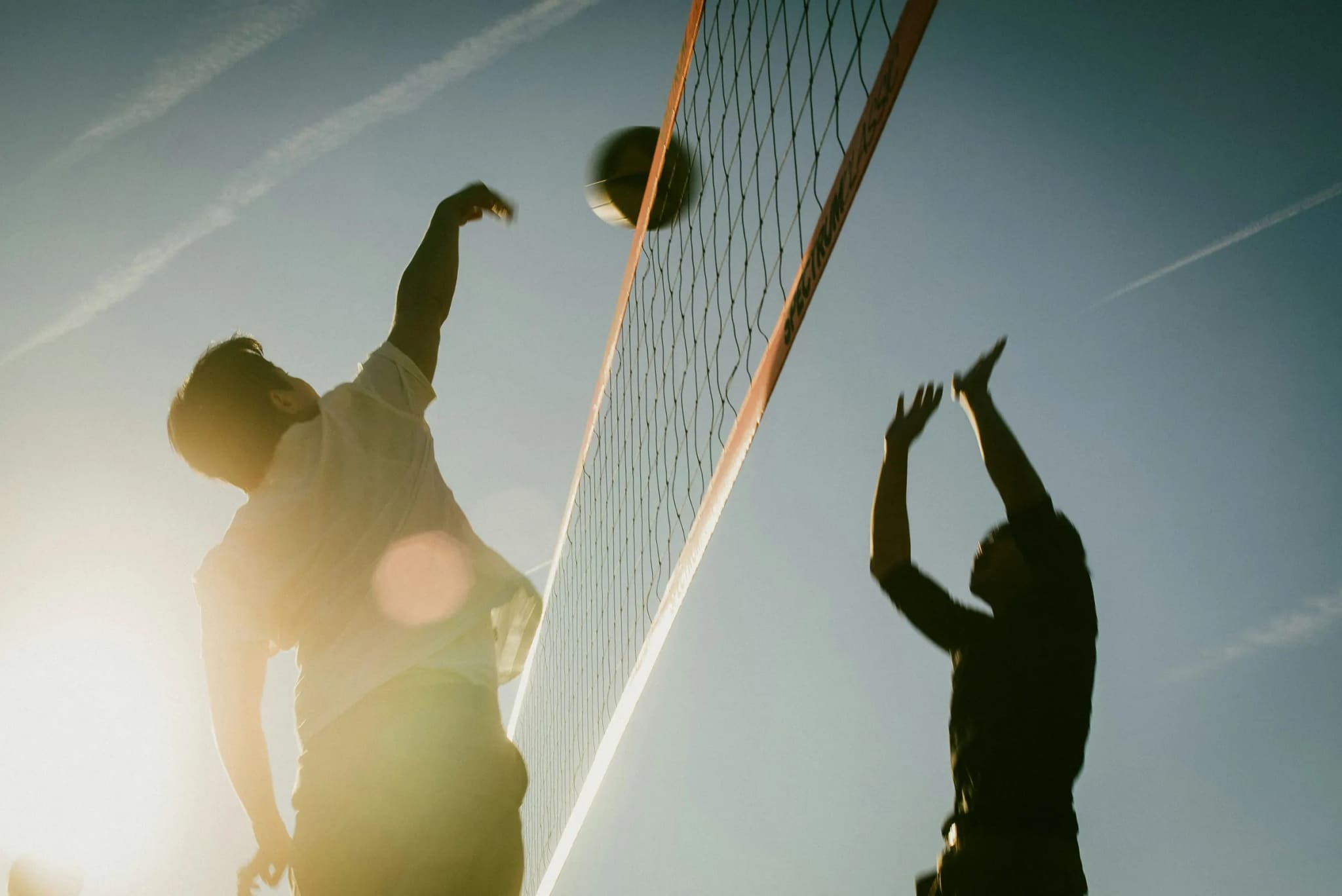 A player spiking the ball over the net against a bright sky, a blocker reaching up on the other side