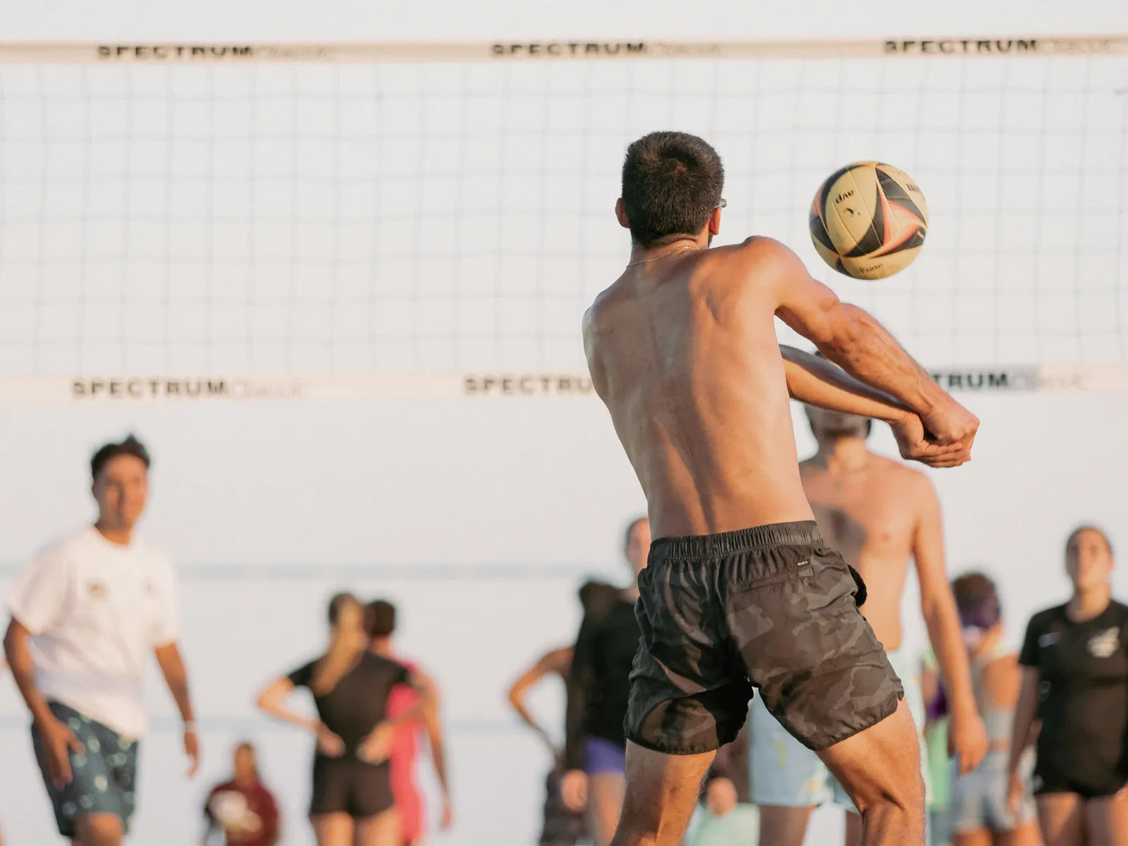A player passing the ball on a beach court at dusk, the rest of the game waiting behind the net