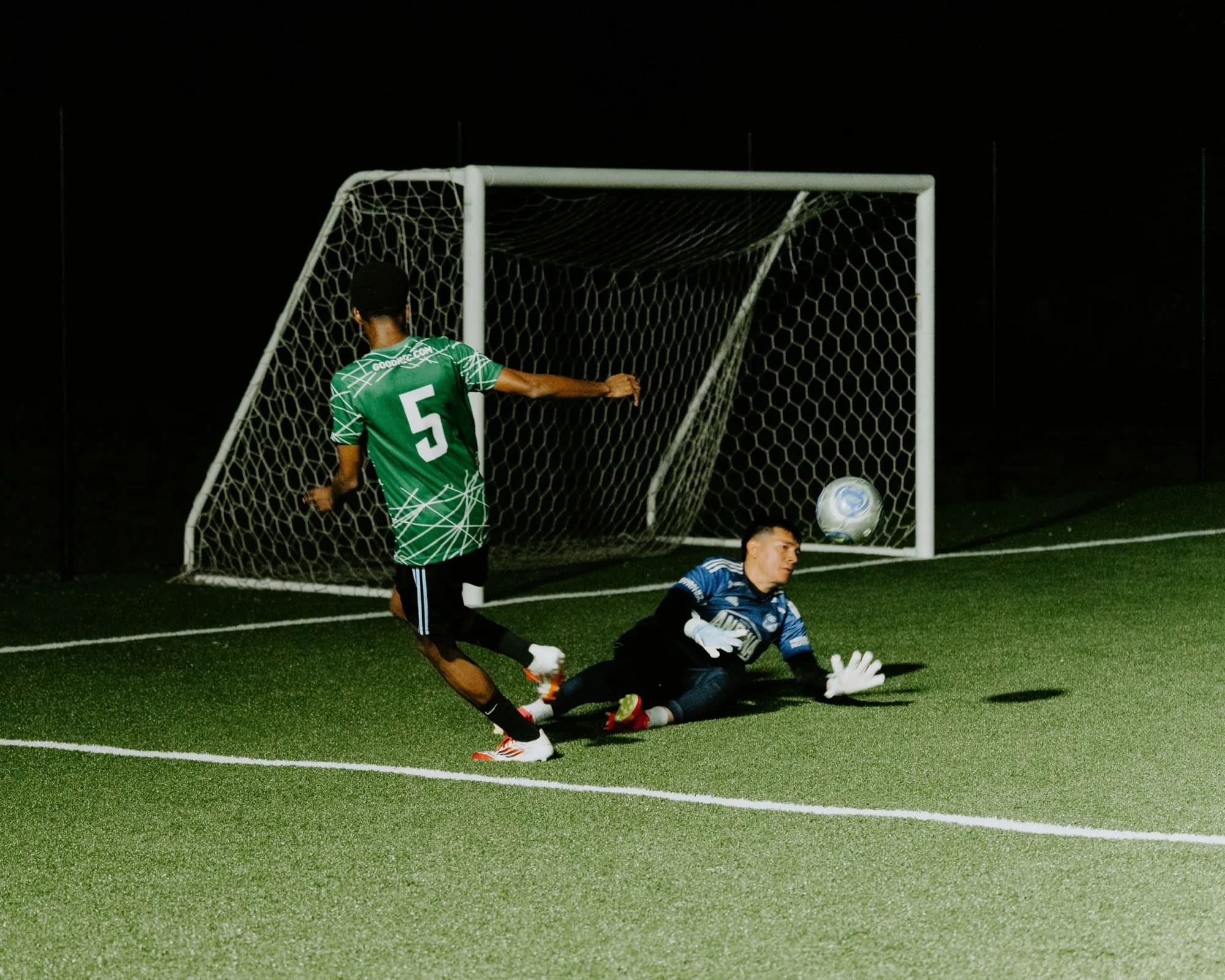 A GoodRec player slotting the ball past a diving goalkeeper under floodlights