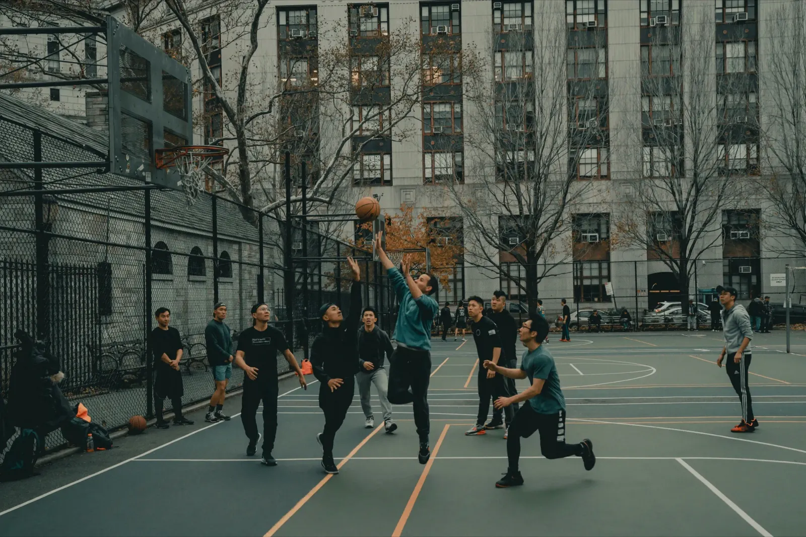 Players on an outdoor city basketball court, one rising for a shot as the others watch the ball