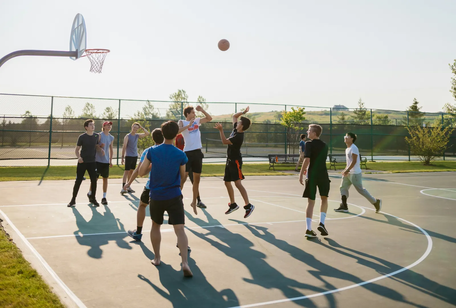 A full pickup game on an outdoor court in the evening sun, a shot in the air and players spread across the key