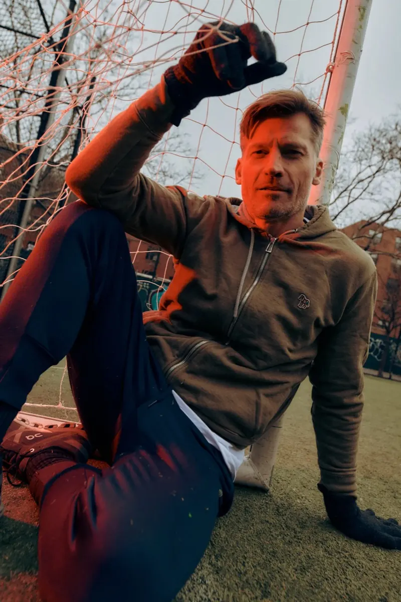 Nikolaj Coster-Waldau sitting against a soccer goal on a city field